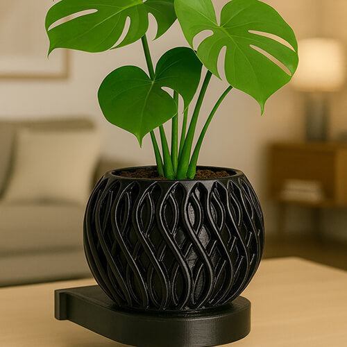A black 3D-printed planter with an intricate interwoven curved pattern, holding a small monstera plant. The pot sits on a matching black base placed on a light wooden table in a softly lit modern living room.