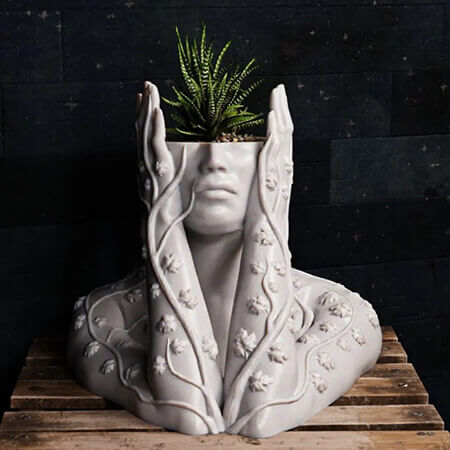 3D printed human face planter with vine detailing and a small green fern plant, artistic sculpture-style design on a wooden surface with dark background.
