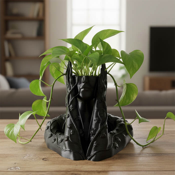 Artistic black 3D printed forest guardian face planter with vines and green trailing plant on wooden table indoors.