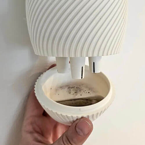 Hand holding the detachable drainage cup of a white 3D-printed hanging planter, showing collected dirty water dripping from the planter’s drainage nozzles.
