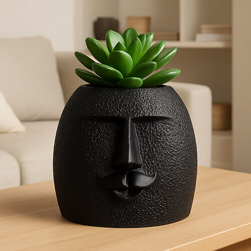 Black textured 3D-printed face planter with a stylized nose and mustache, holding a small green succulent, placed on a wooden table in a modern living room.