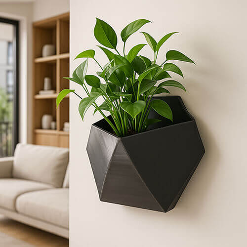 Black geometric 3D-printed wall planter with sharp faceted design, holding a leafy green plant, mounted on an indoor wall in a modern living room.