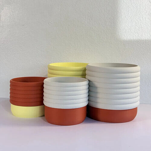 Stacked set of ribbed self-watering planters in white, terracotta, and pastel yellow colors arranged against a light wall, with no plants inside.