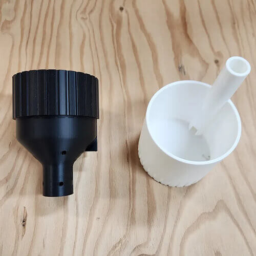 Disassembled self-watering planter shown on a wooden surface, featuring a black upper funnel component with drainage holes and a white lower container with an attached vertical water-fill tube.